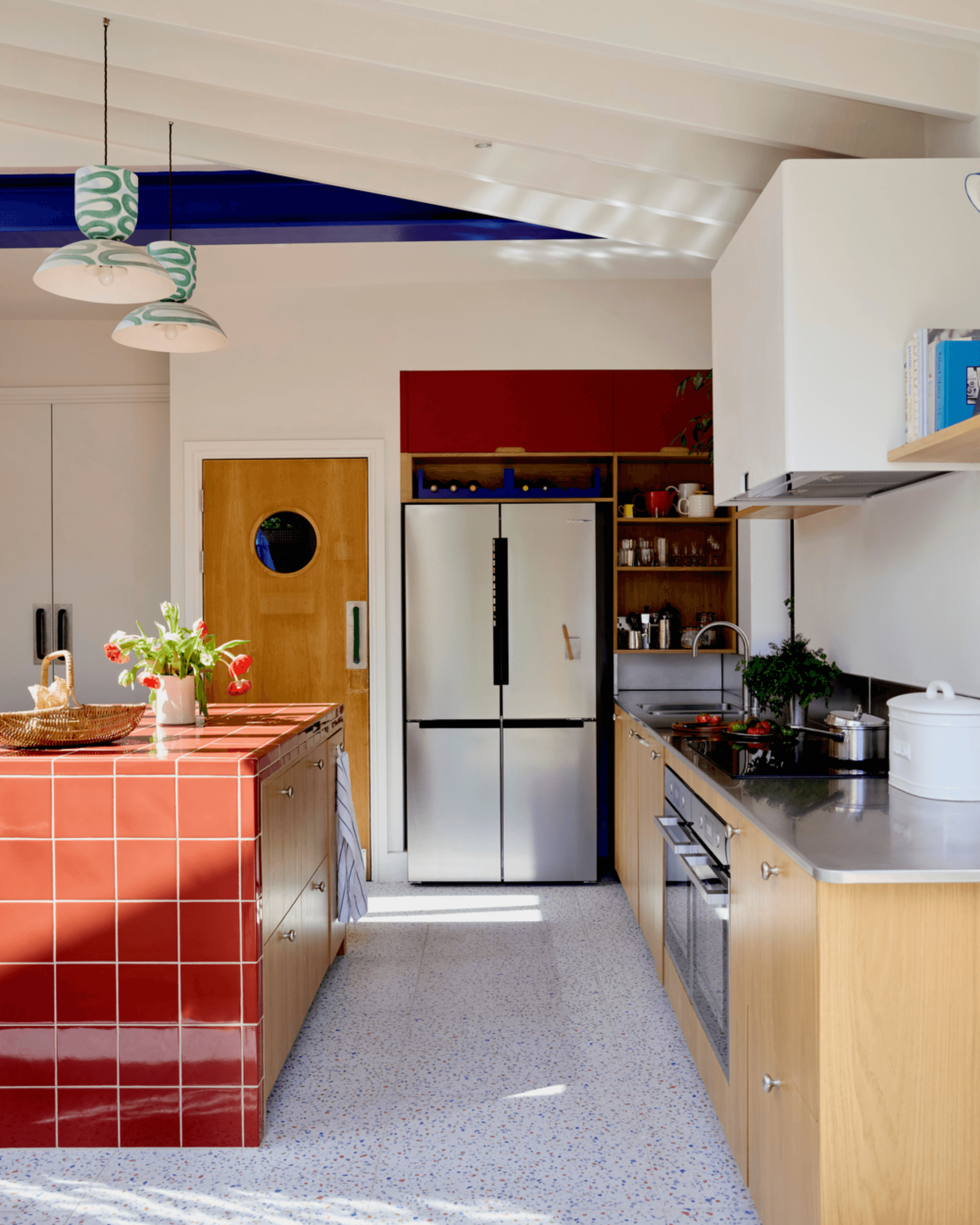 Wide angle of a vibrant kitchen featuring red tiled island, oak veneer and laminate fronts, and stainless steel worktop.