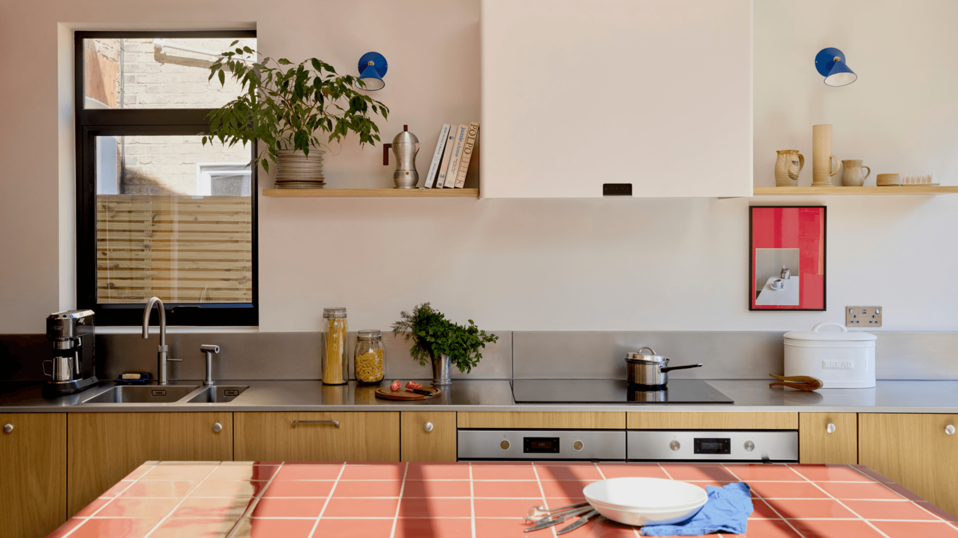 Wide angle photo of a kitchen featuring red tiled island, oak veneer fronts and stainless steel worktop.